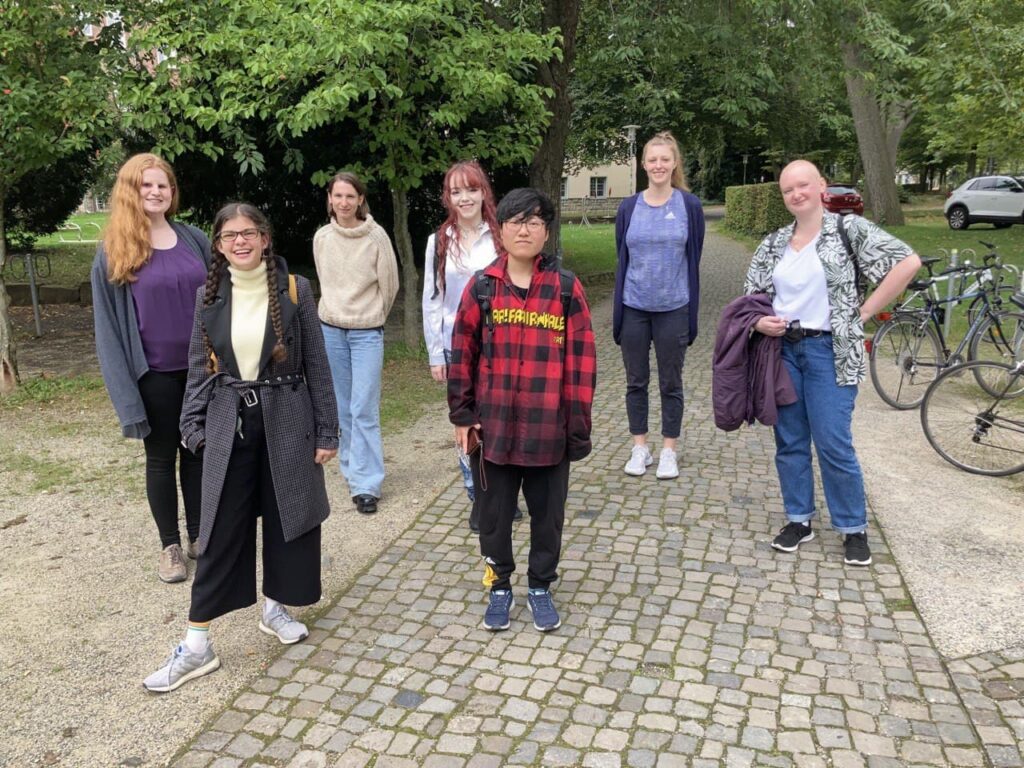 Photo of seven people on campus in front of some trees. The people are smiling and standig in slightly offset positions in social distancing approved distances.